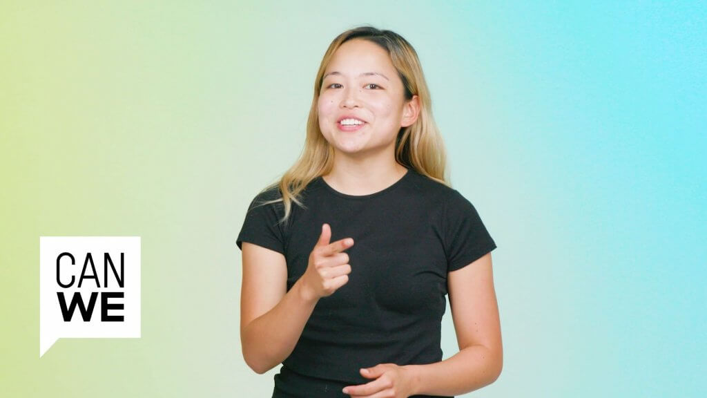 A young person from a South or Southeast Asian background, with medium toned skin and long brown and blonde hair half-smiles at the camera. One hand is raised, pointing towards the audience.
